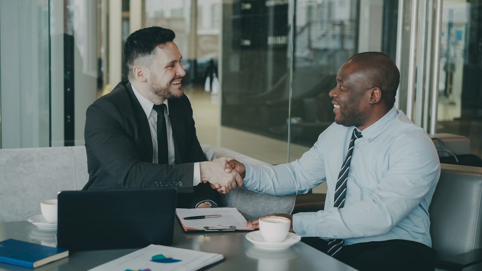 Two individuals in formal attire shaking hands and smiling in a professional meeting setting.